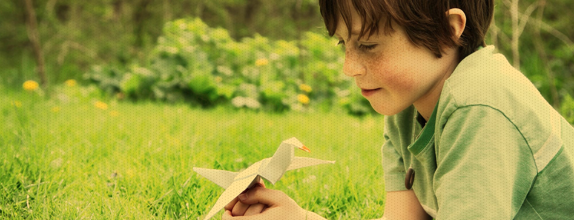 Little boy laying in grass, smiling and holding an origami bird made of paper