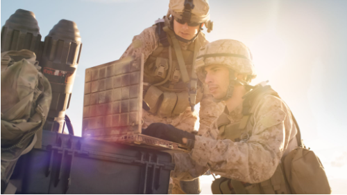 Two military service members in desert camouflage and tactical helmets operate a ruggedized laptop resting on a protective storage case.