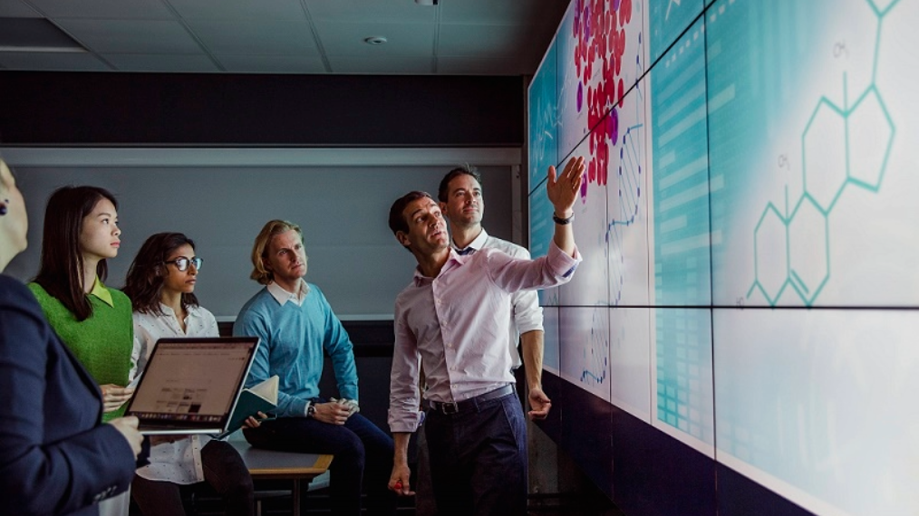 Six people standing in a room looking at various graphs on a big screen , while one of the six people points to it