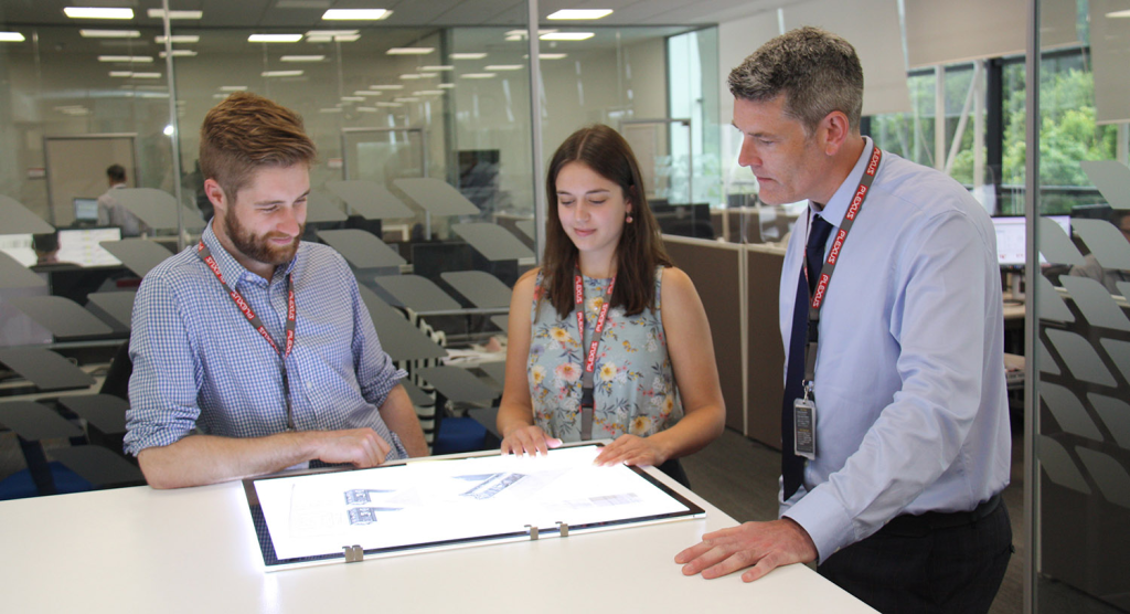 Three engineering professionals collaborate around a large, illuminated light table to closely review technical design blueprints. The team, wearing business casual attire and company lanyards, is focused on the schematics within a modern, open-plan office environment featuring glass walls.