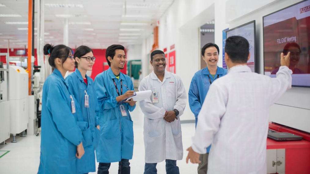 A diverse team of six manufacturing professionals in blue and white smocks gathers for a stand-up meeting in a bright Plexus facility. A team leader stands at the front, gesturing toward a digital presentation screen, while the group listens attentively and smiles, illustrating cross-functional collaboration.