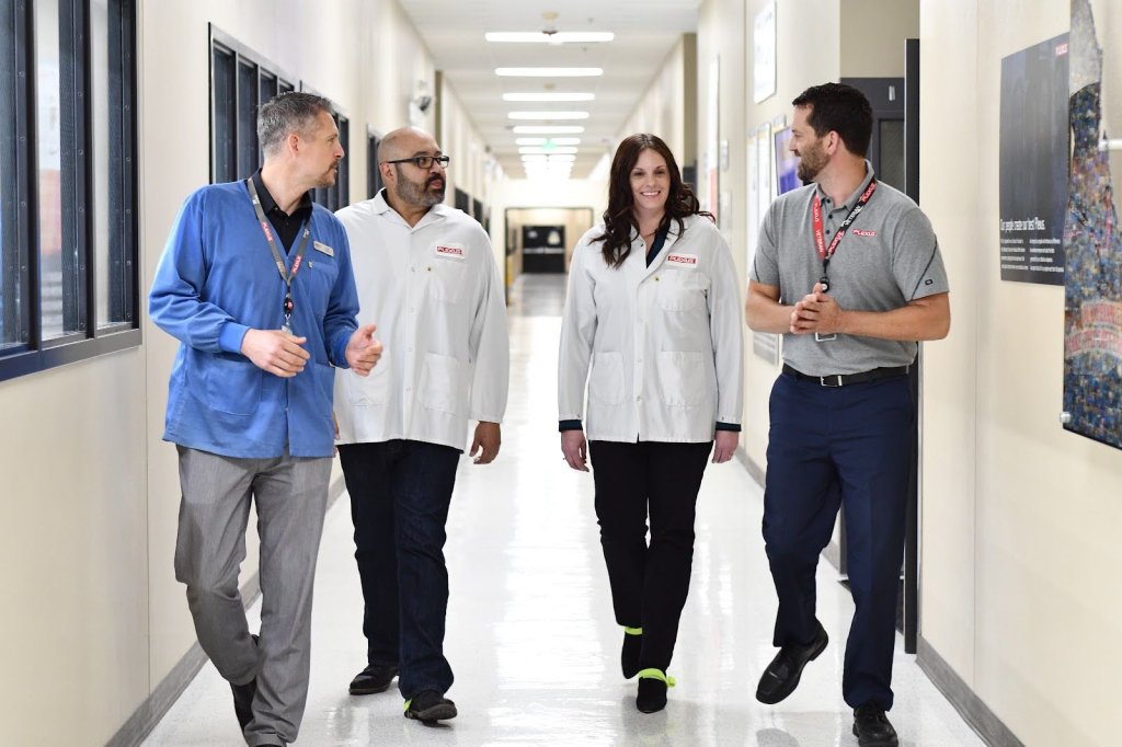 Four professionals—including individuals in blue and white lab coats—walk side-by-side down a bright hallway in a Plexus facility, engaged in active conversation. The group is smiling and talking, illustrating the strong relationships and continuous communication between transition managers and manufacturing teams.
