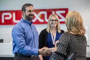A smiling man in a blue shirt is shaking hands with a woman. Another woman with blonde hair and glasses is smiling in the background. The Plexus logo is on a wall behind them.