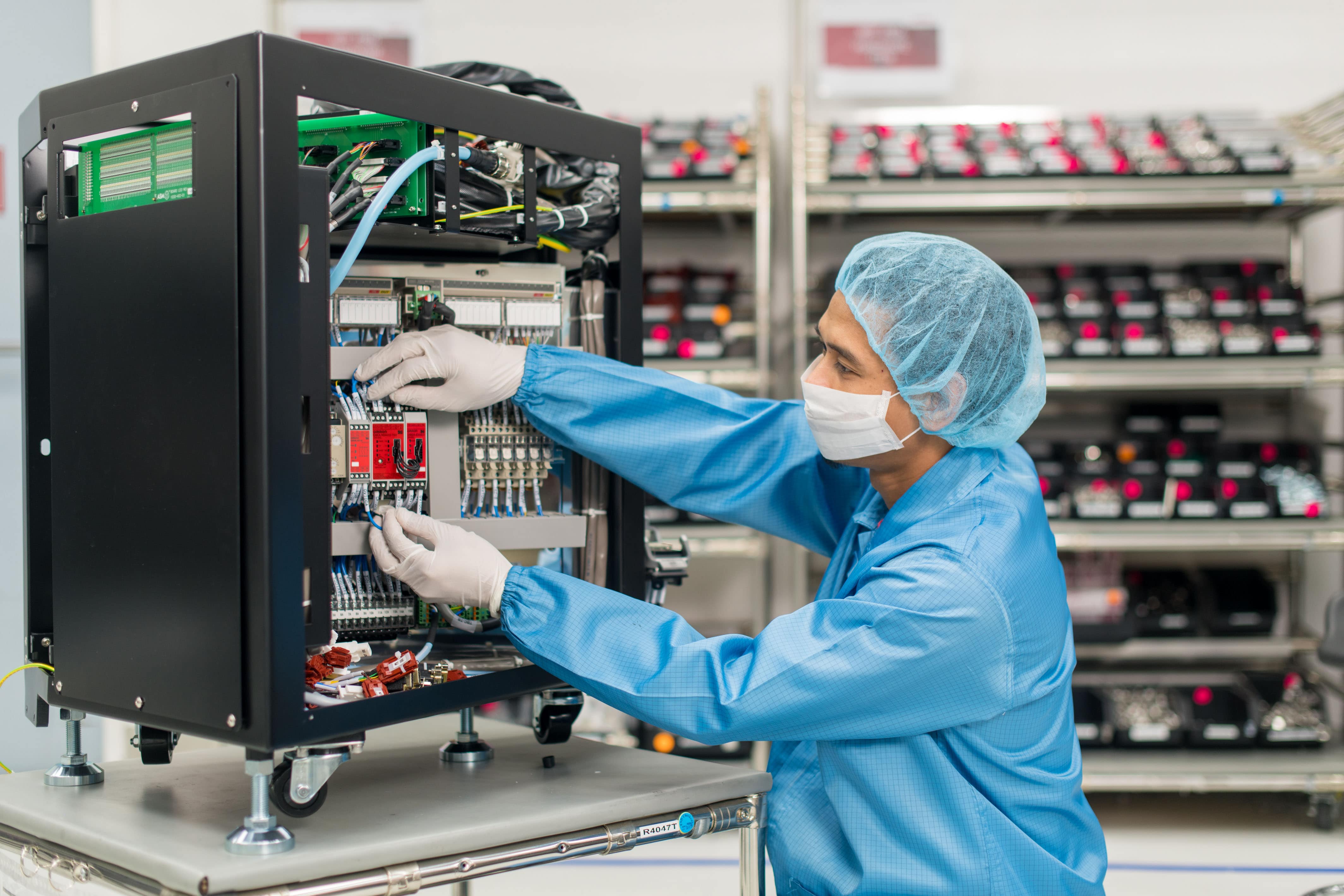 A technician in cleanroom attire assembles or inspects electrical components inside a control unit in a high-tech manufacturing environment.