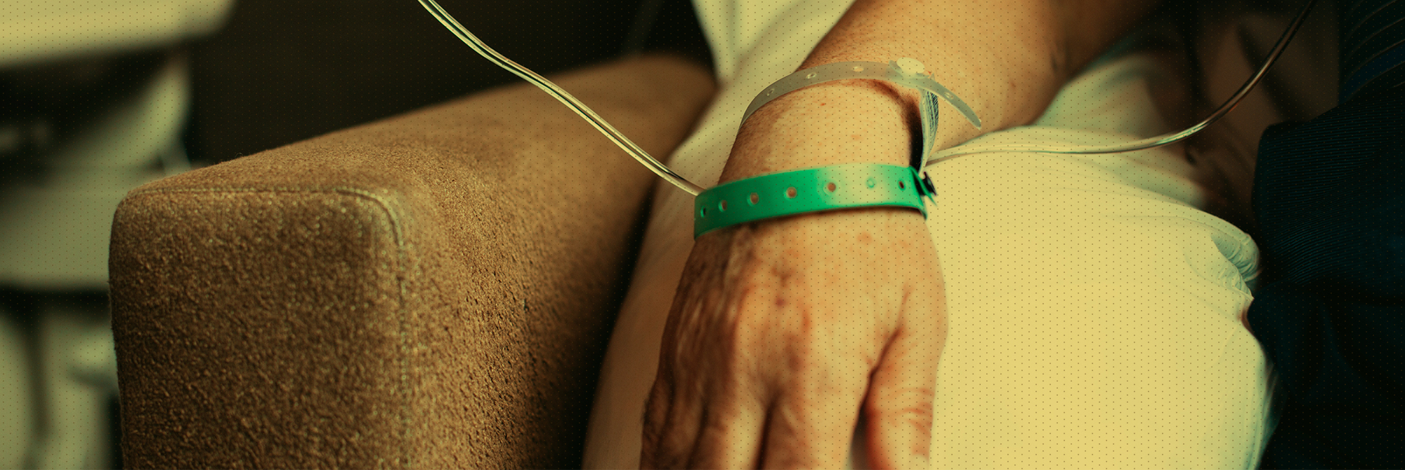 A close-up of a person's arm with a small tube and a green medical wristband, resting on a beige armchair