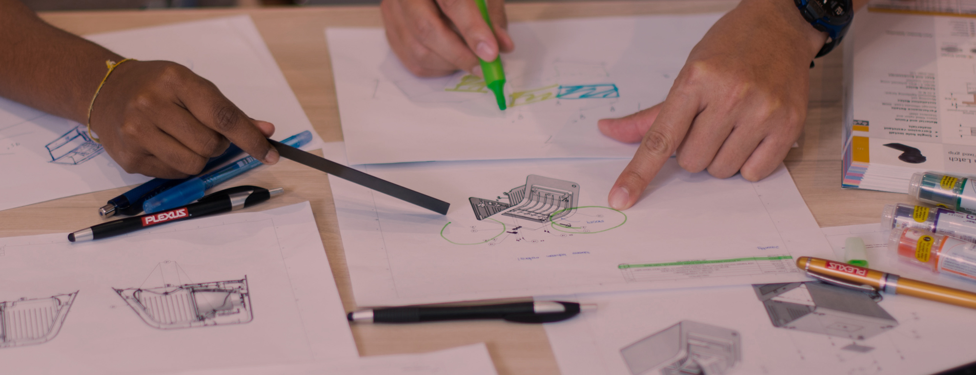 Two people collaborate over technical drawings and product design sketches, using markers and pens to highlight and annotate engineering details.