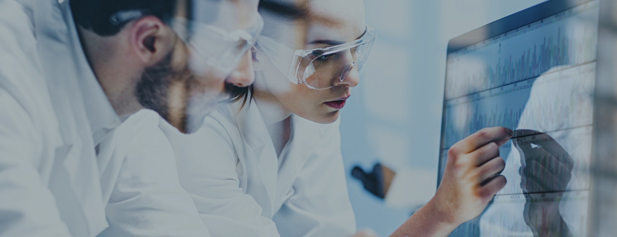 Two scientists wearing lab coats and safety goggles analyze complex data visualizations on a large digital screen in a laboratory setting.