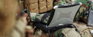 A close-up view shows a soldier in camouflage and tactical gear using a ruggedized laptop in a field environment.