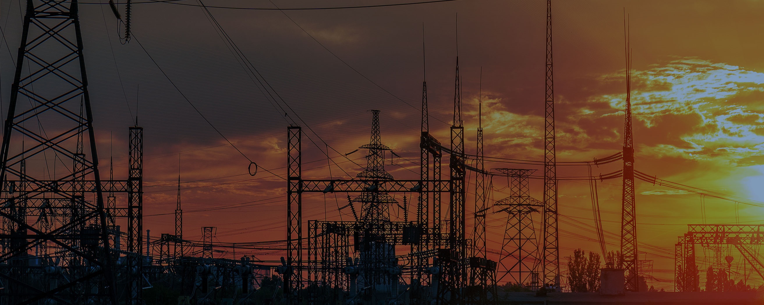 High-voltage power lines and transmission towers are silhouetted against a dramatic sunset sky at an industrial substation.