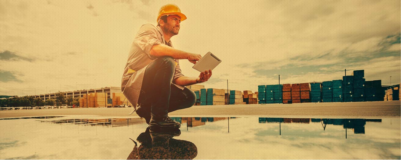 A male construction worker or engineer in a yellow hard hat and work clothes squats next to a puddle, holding a tablet and looking towards a large outdoor storage area filled with stacked containers or materials, with the sky and containers reflected in the puddle