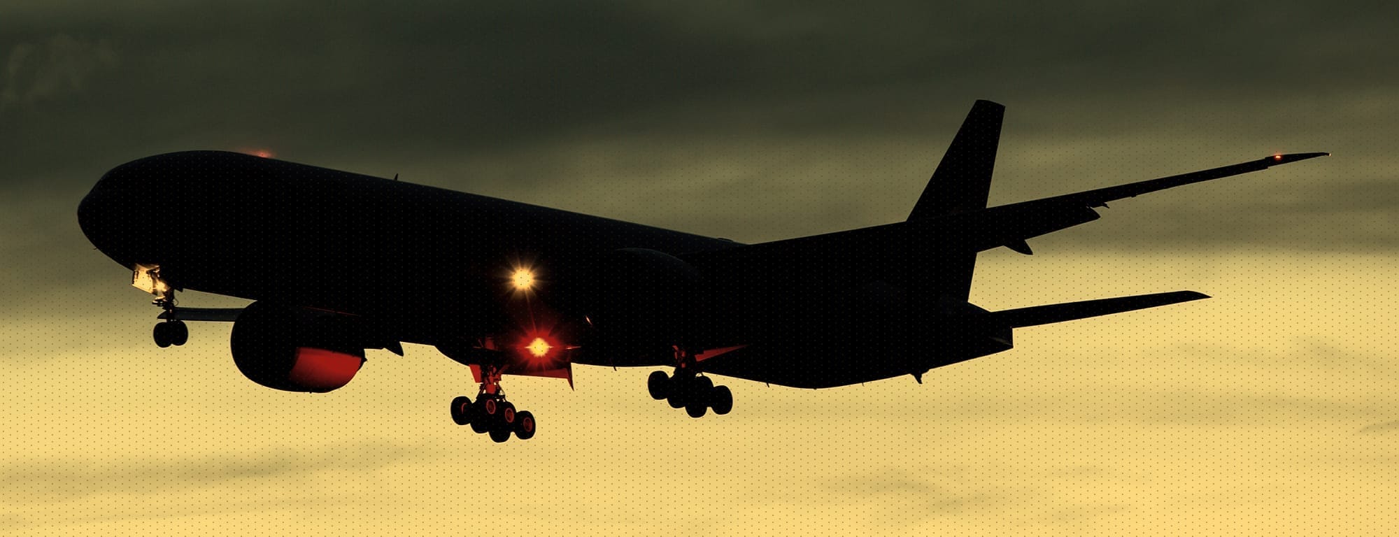 A large airplane, silhouetted against a dark, cloudy sky, is in mid-air with its landing gear down and illuminated by bright orange lights on its underside and wingtips