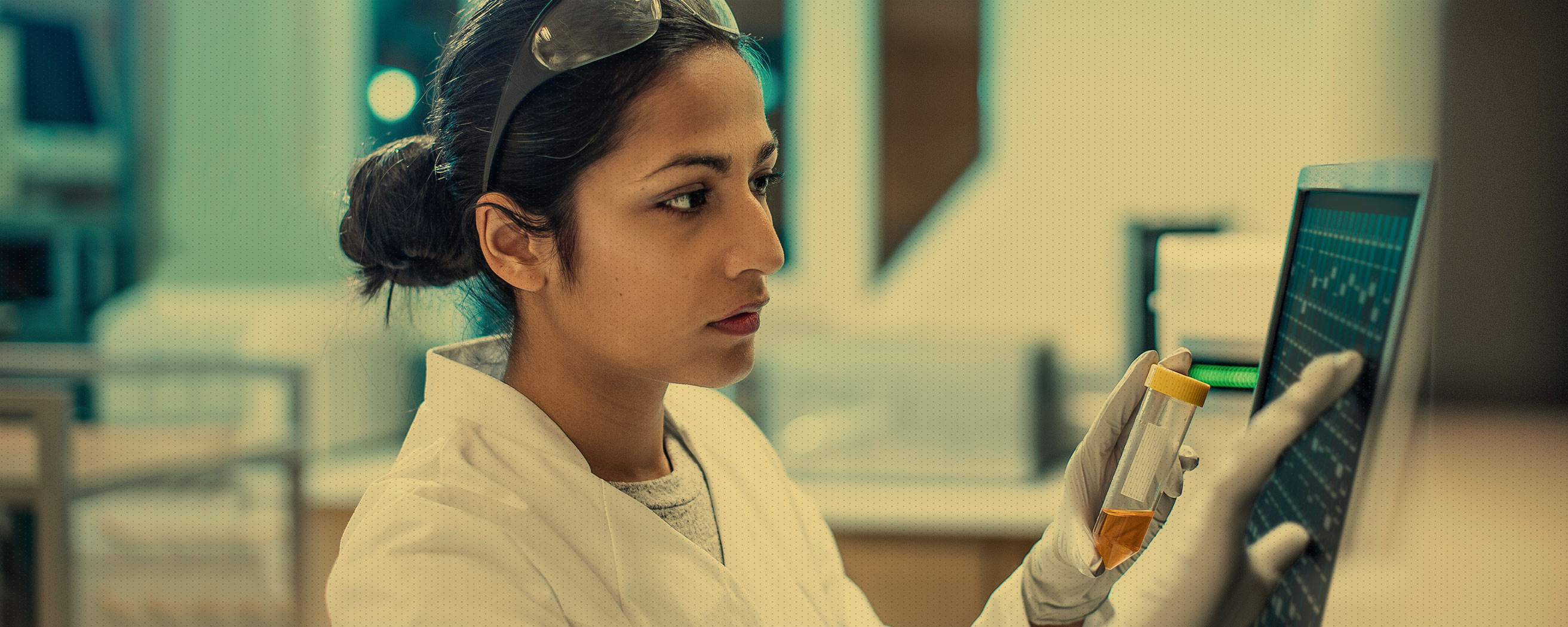 A scientist in a lab coat and safety glasses, wearing gloves, looks at a laptop screen with graphs while holding a test tube filled with liquid