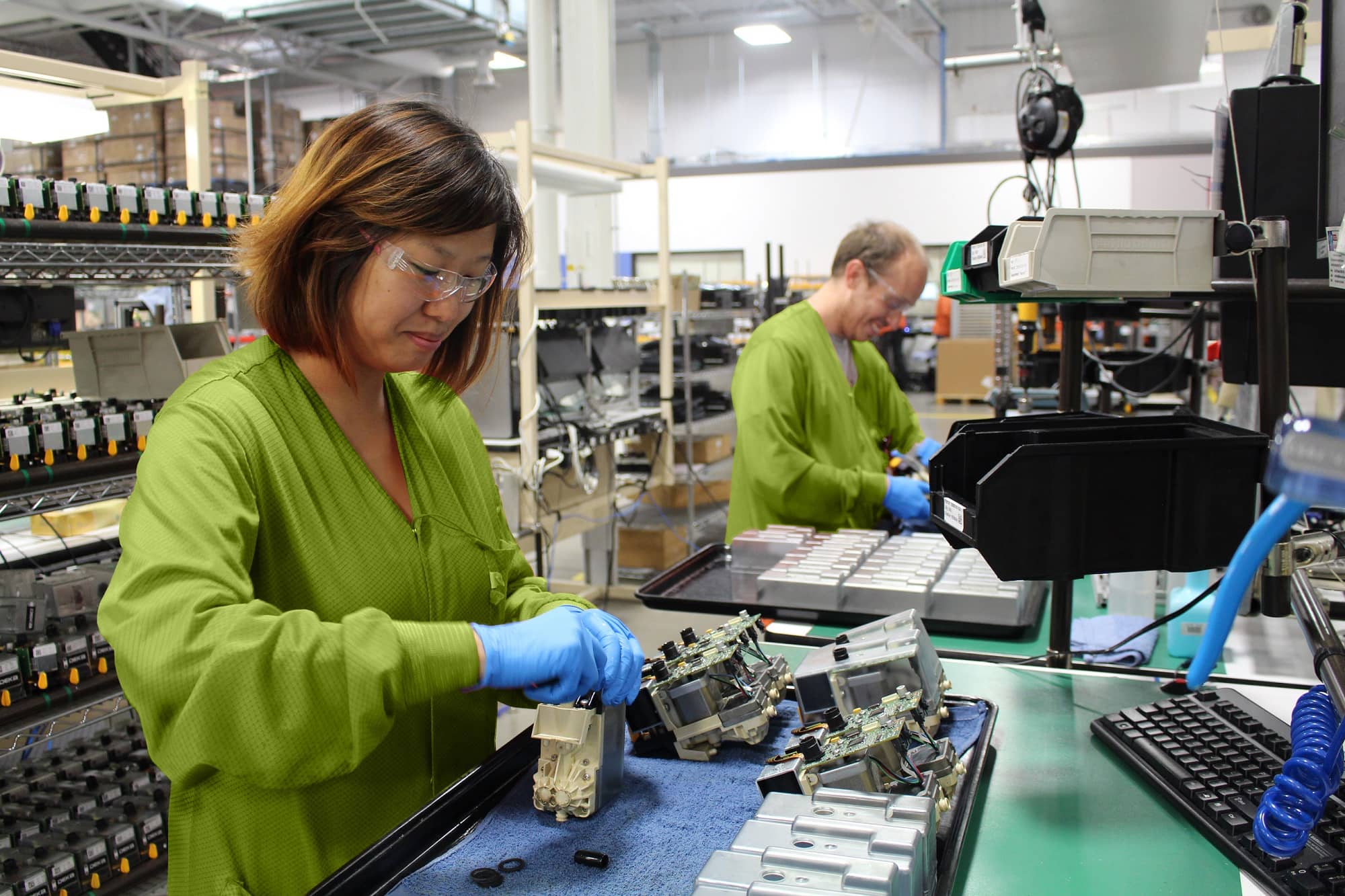 Two workers in green lab coats and safety gear assemble electronic components at a workstation in a modern manufacturing facility.