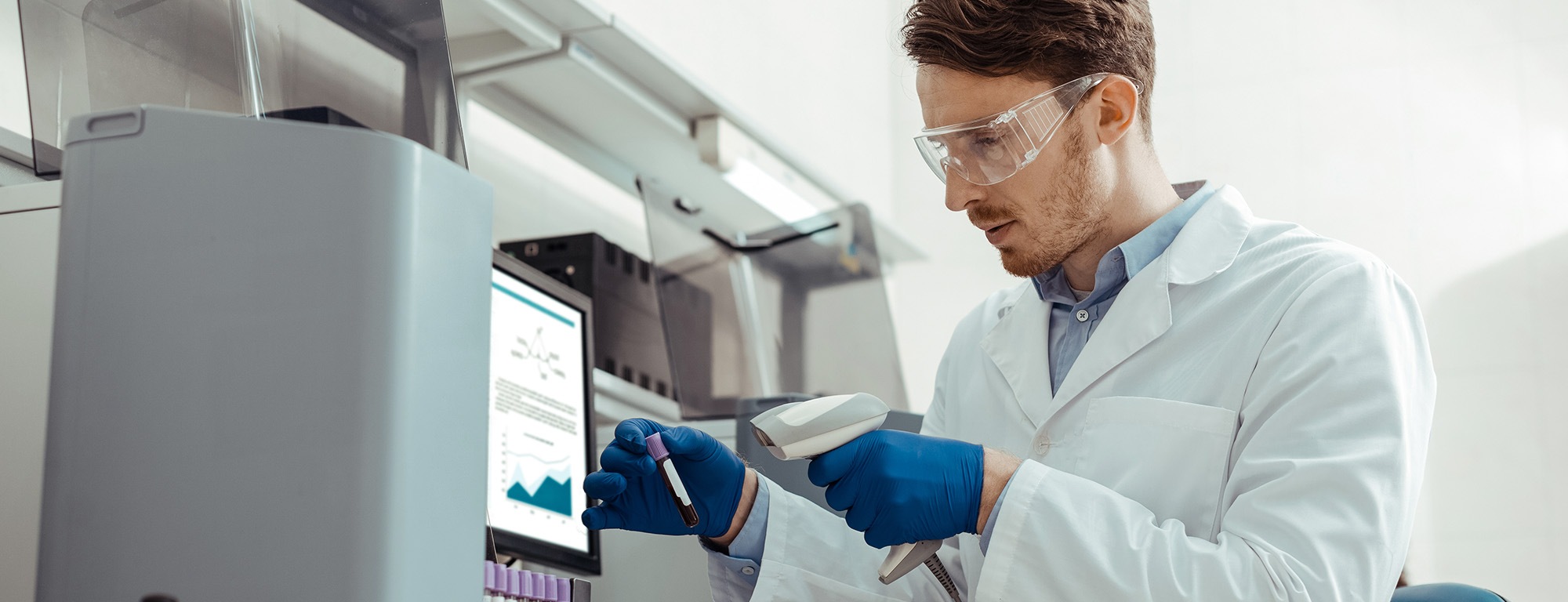 A scientist in a lab coat and safety goggles analyzes a blood sample using a barcode scanner, with data displayed on a nearby monitor.