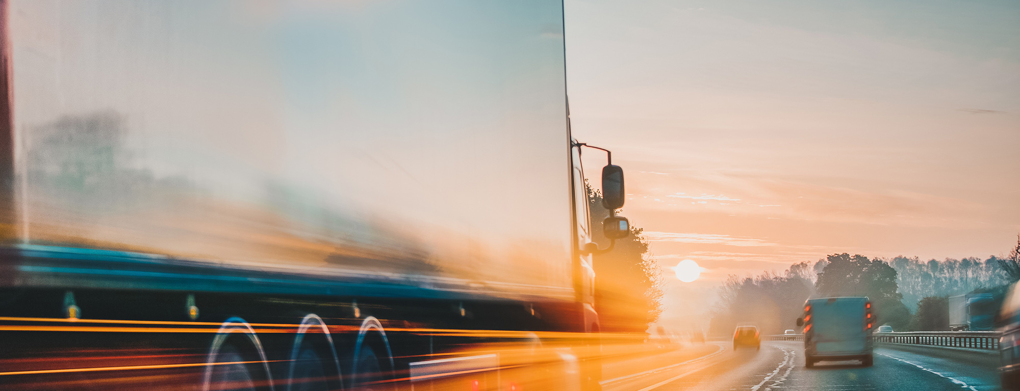 A motion-blurred truck speeds down a highway at sunrise, surrounded by other vehicles and framed by a glowing horizon.