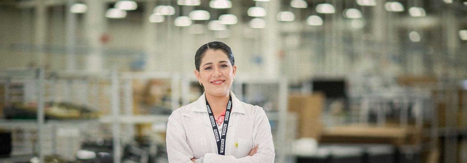 A smiling woman wearing a lab coat and ID badge stands confidently in a brightly lit manufacturing or assembly facility.