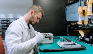 A male technician wearing a white lab coat and gloves sits at a desk, carefully working on the internal components of a small white electronic device.