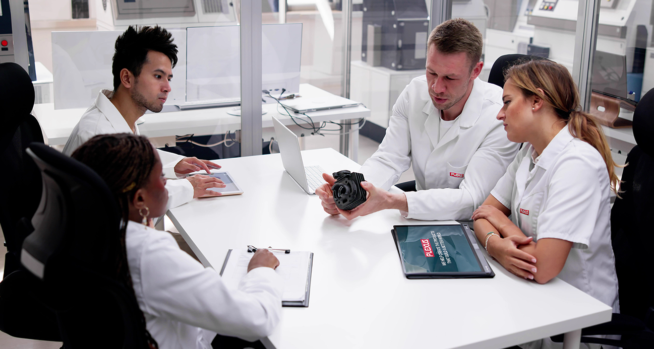 Four professionals wearing white lab coats branded with the "Plexus" logo sit around a white table in a modern laboratory setting. A man in the center holds a black mechanical component, discussing it with his three colleagues who are listening attentively. On the table are a laptop, a clipboard, and a tablet displaying the Plexus logo and the text, "Help create the products that build a better world."