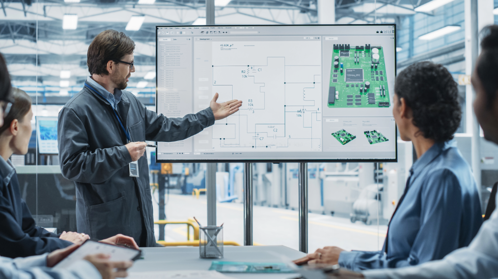 A lead engineer presenting a complex printed circuit board (PCB) schematic and 3D assembly model on a large digital display to a team of seated colleagues. This collaboration illustrates Plexus’ role as a strategic partner, providing specialized systems engineering and "copy-exact" manufacturing for semiconductor OEMs to overcome talent shortages and accelerate innovation.