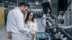 Two professionals, a man and a woman, are wearing white lab coats and working together in a high-tech manufacturing facility. They are leaning over a workstation where a green circuit board is mounted on a testing fixture. The man is pointing to a specific component on the board while the woman looks on attentively. Both are wearing glasses, and their lab coats feature a red "PLEXUS" logo. The background shows a spacious, well-lit industrial environment with various pieces of technical equipment.