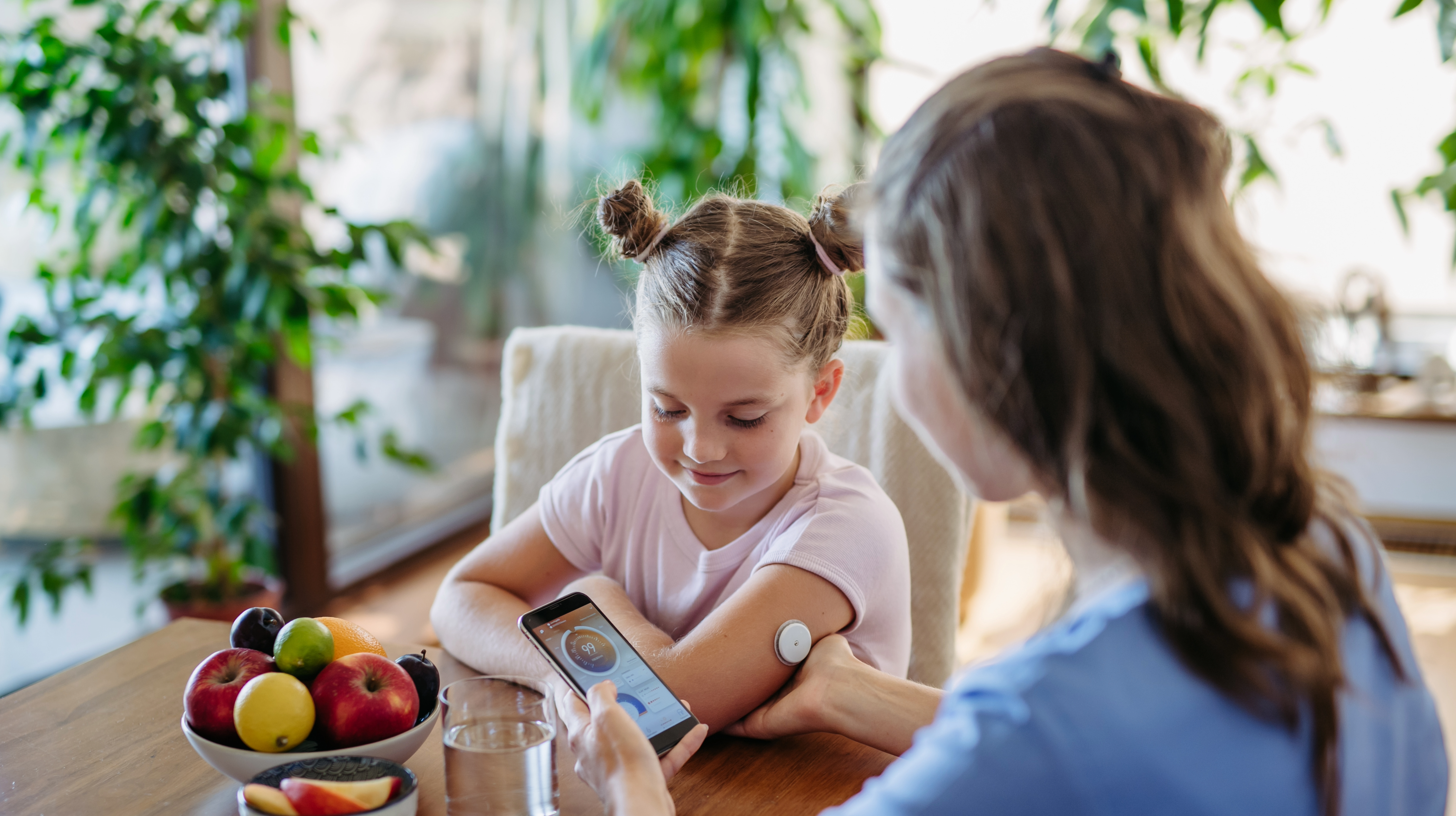 A young girl with a glucose monitor on her arm watches as a woman holds a smartphone displaying a monitoring app. They are sitting at a wooden table with a bowl of fruit and a glass of water.