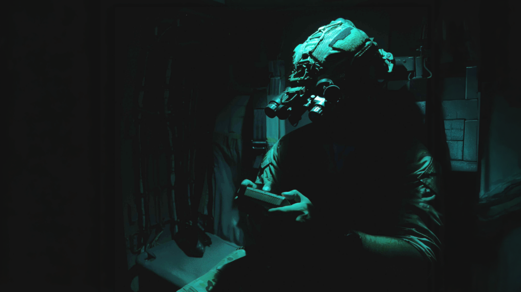 A soldier in a dark environment, illuminated by a low-profile green light source. The soldier is wearing a tactical helmet equipped with quad-lens night vision goggles (NVGs) and appears to be holding or operating a handheld device, such as a controller or tablet.