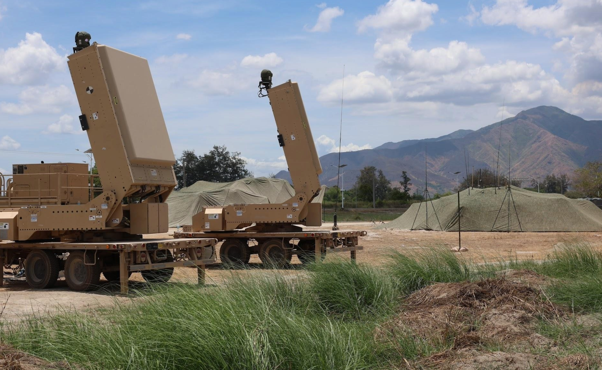 Two military counter-UAS radar units parked in a grassy field with a camouflaged command tent and mountains in the background under a cloudy sky.