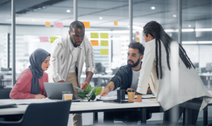 Ein vielfältiges Team aus vier Fachleuten hat sich um einen großen weißen Tisch in einem hellen, modernen Büro versammelt. Eine Frau mit Hijab sitzt vor einem Laptop, während ein Mann in einem gestreiften Hemd neben ihr steht und auf den Bildschirm schaut. Ein weiterer Mann zeigt auf Dokumente auf dem Tisch, während er mit einer Frau mit langen Zöpfen spricht, die ihm gegenüber steht. Im Hintergrund ist eine Glaswand mit bunten Haftnotizen bedeckt, was auf eine Brainstorming- oder Projektplanungssitzung hindeutet.