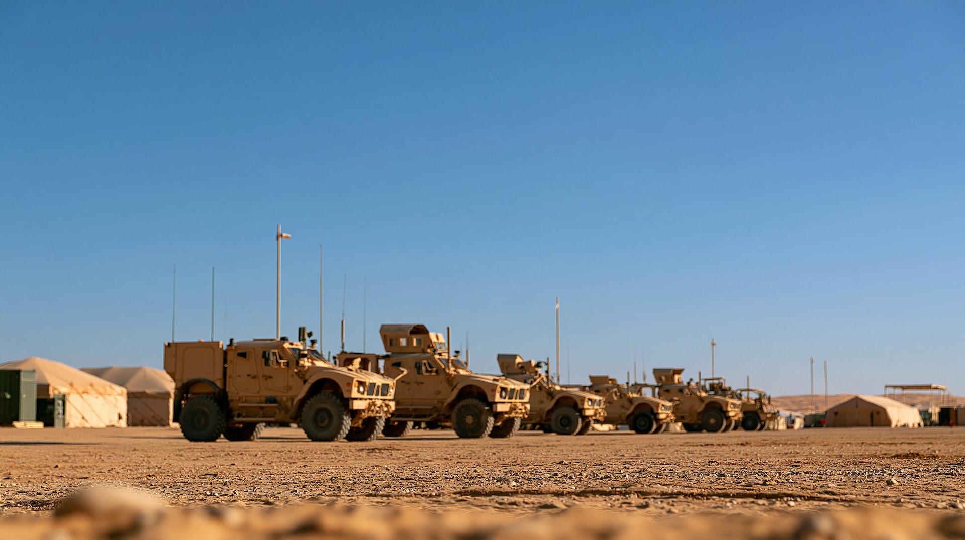A line of tan military armored vehicles parked at a desert base under a clear blue sky.
