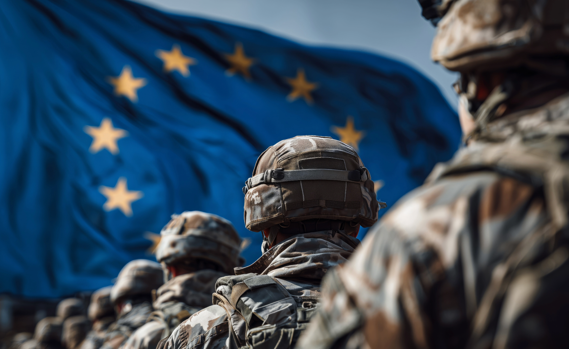 A close-up view from behind a line of European soldiers in military uniforms and combat helmets, standing in formation in front of a large, blurred European Union flag with yellow stars flying against a blue sky.