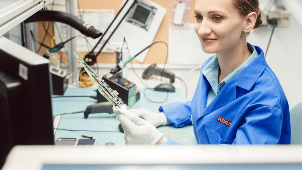 A Plexus electronics technician wearing a blue smock and white protective gloves, smiles while holding and inspecting a green printed circuit board assembly at a laboratory test station.