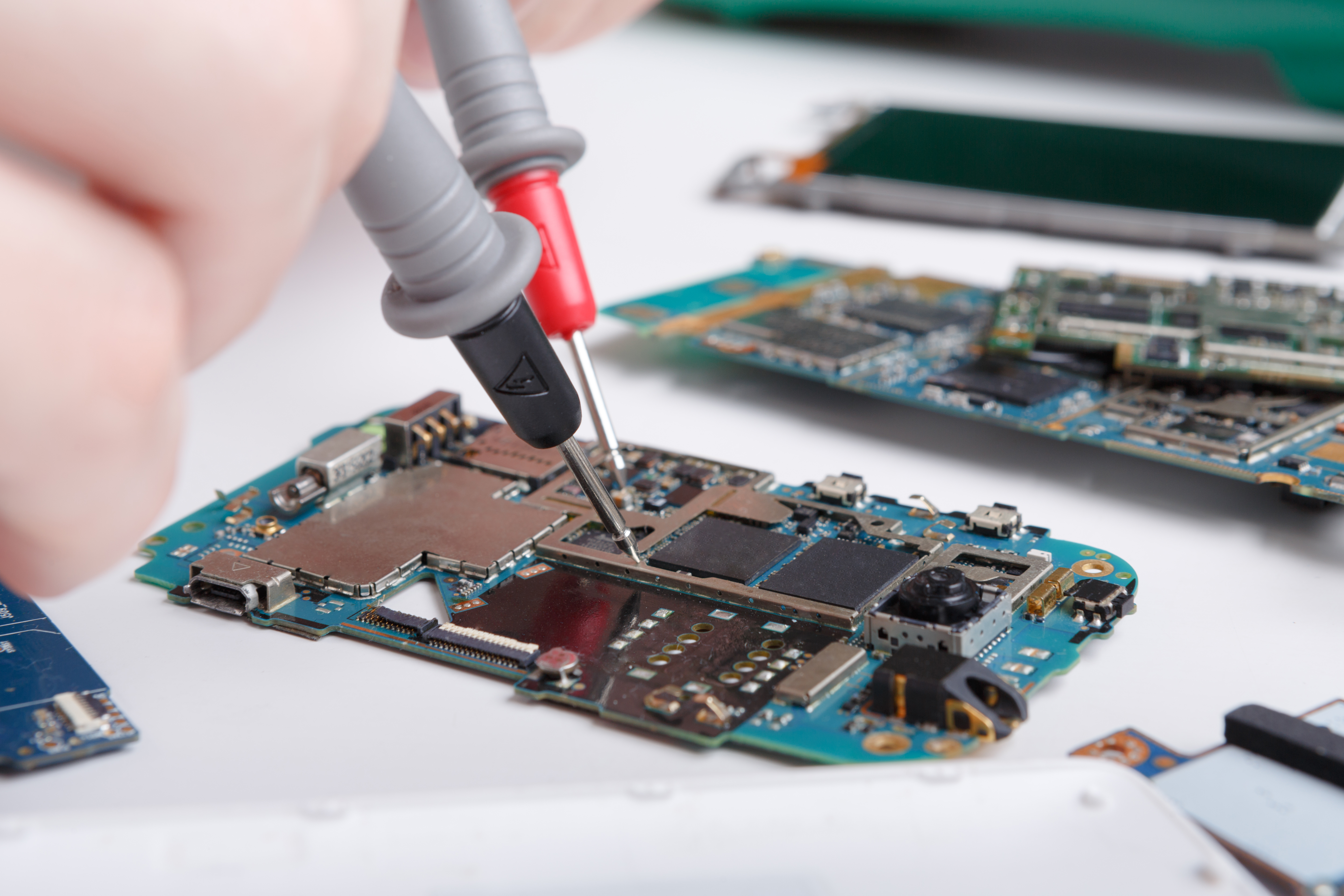 A close-up view of an electronics technician using diagnostic test probes to check an exposed printed circuit board assembly (PCBA) on a workstation, with disassembled hardware components visible in the background.