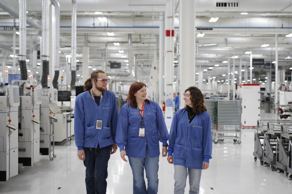 Three workers wearing blue protective lab coats and ID badges smile and talk as they walk together across a bright, modern manufacturing floor. 
