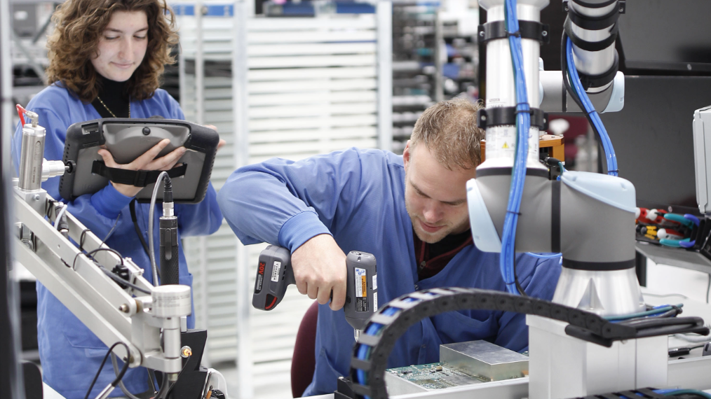 Two operators in blue coats work at a workstation, one using a power driver on an assembly next to a robotic arm while the other monitors a tablet.